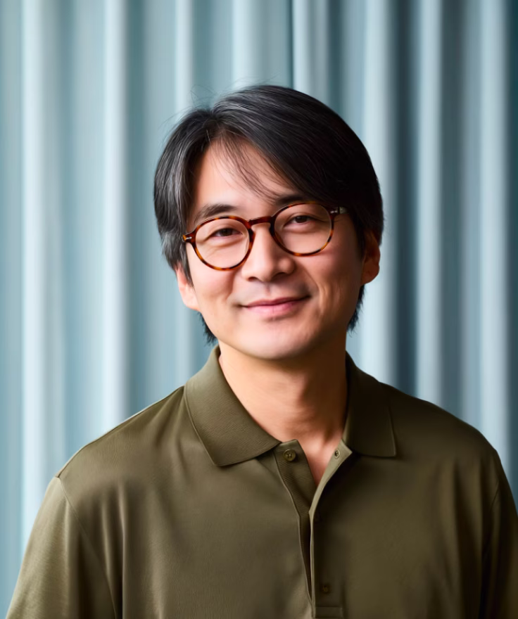 Smiling man with black hair and tortoiseshell glasses wearing an olive green polo shirt against a light blue striped background.