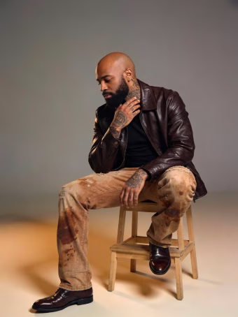 Bearded man with tattoos wearing a brown leather jacket and tan pants, sitting on a wooden stool against a gray background.